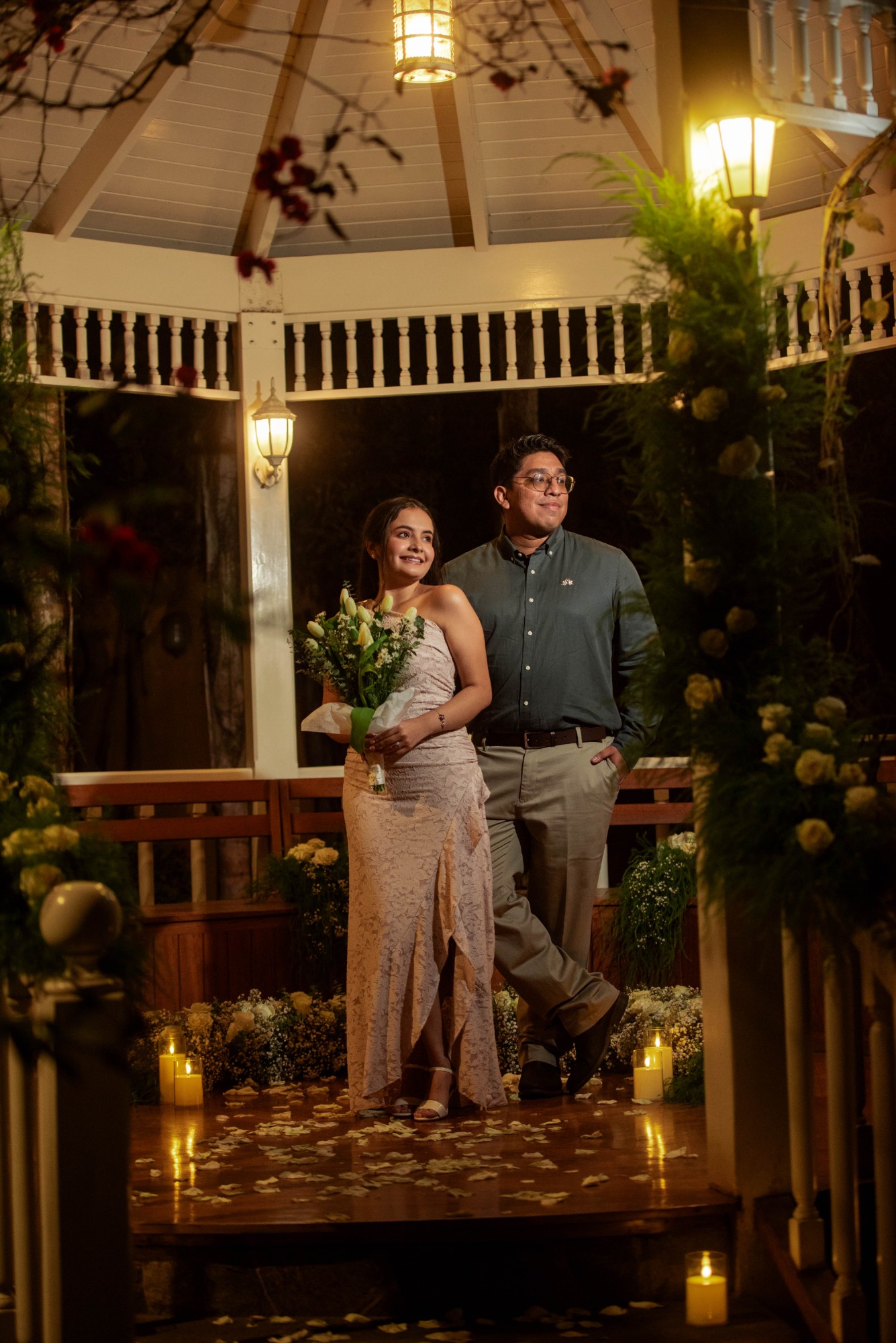 Angie en la glorieta iluminada e Iván sentado en el jardín, de noche.