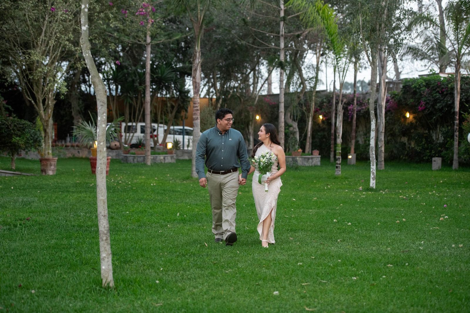 Iván y Angie caminan de la mano por el jardín y se miran; ella lleva un ramo de tulipanes blancos.